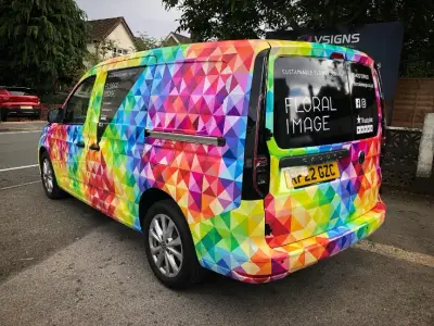 Rear quarter of Floral Image VW Caddy van in colourful geometric full wrap, with V Signs board behind
