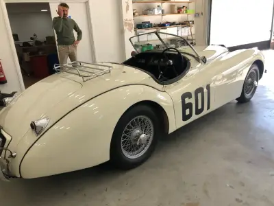 Cream Jaguar XK120 roadster with black 601 race-number graphic, rear-side view in workshop