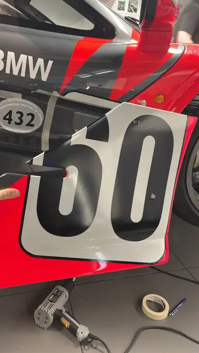 Applying black number 60 door panel to red and grey BMW McLaren race livery, heat gun in use