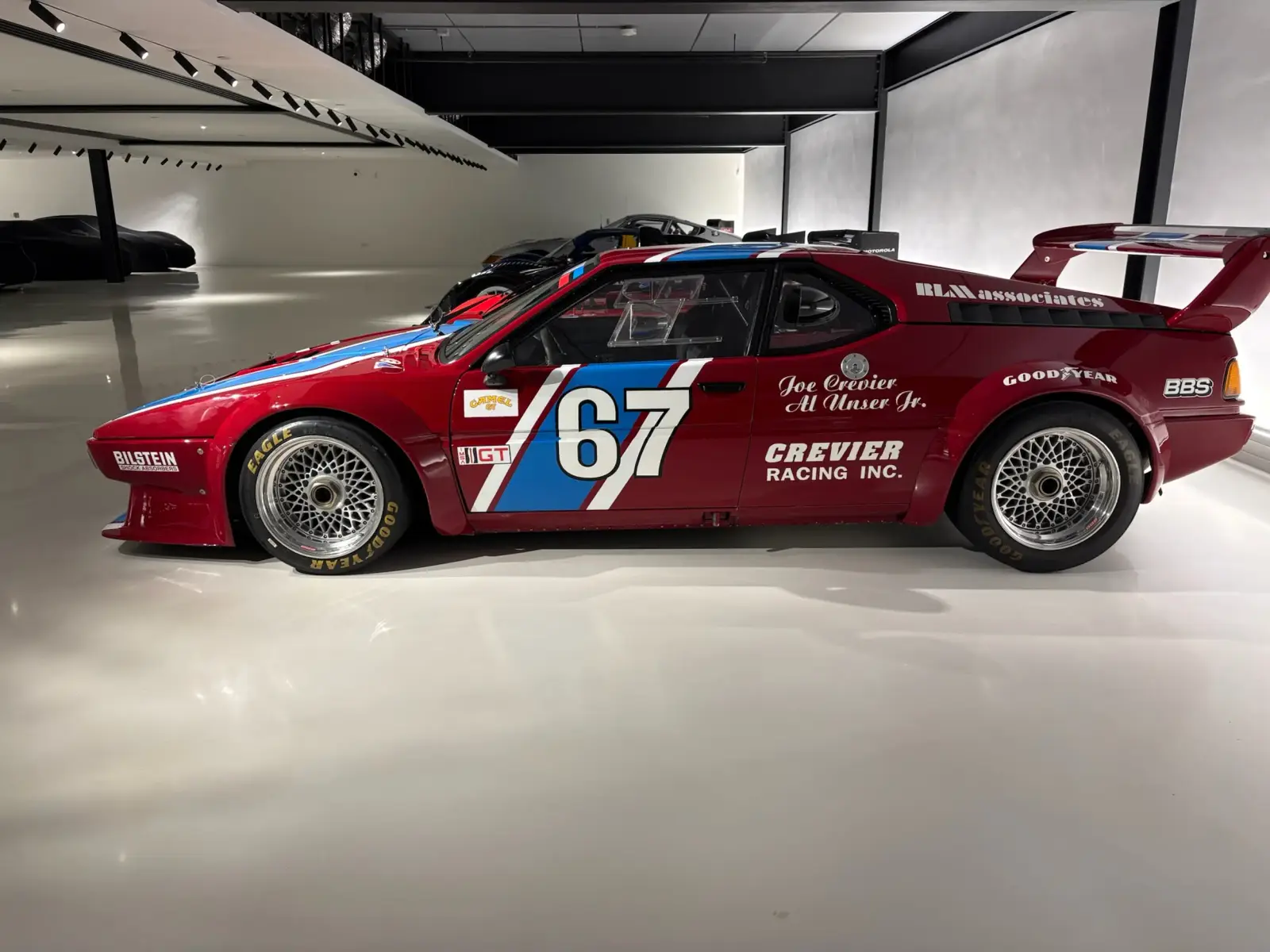 Red BMW M1 Procar race car, number 67, Crevier Racing livery with blue and white stripe, side profile in a showroom