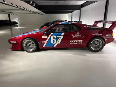 Red BMW M1 Procar race car, number 67, Crevier Racing livery with blue and white stripe, side profile in a showroom