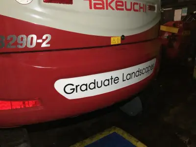 White 'Graduate Landscapes' magnetic sign on the red rear panel of a Takeuchi excavator
