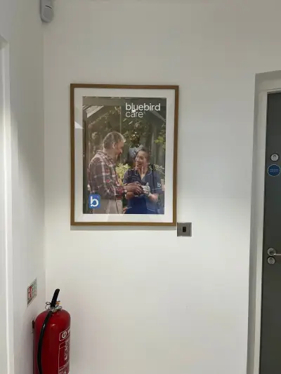 Wood-framed Bluebird Care poster of a smiling carer and gardener hung in a hallway