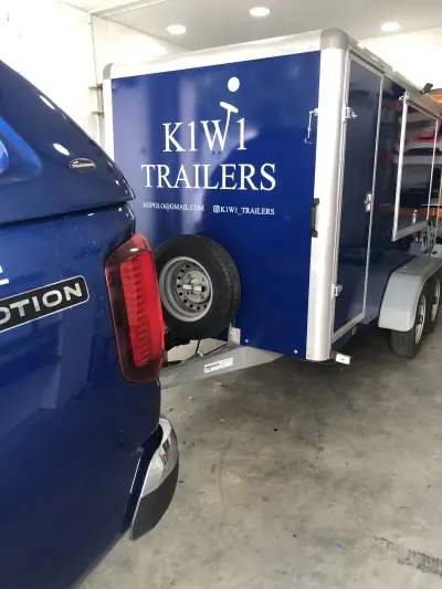 Navy blue K1W1 Trailers enclosed trailer, rear corner with spare wheel and white graphics, beside a blue pickup