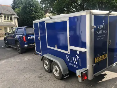 Navy blue K1W1 Trailers enclosed trailer towed behind a blue Kendall crew-cab pickup on a street