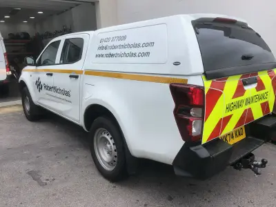 White Isuzu pickup with Robert Nicholas civil engineering graphics and Highway Maintenance chevron rear