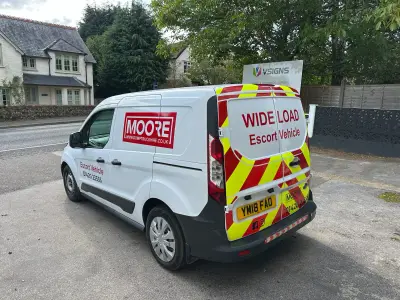 White Ford Transit Connect van with red Moore logo and red-yellow WIDE LOAD escort chevrons