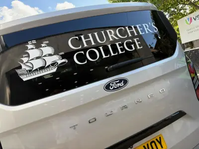 Rear window of silver Ford Tourneo with white Churcher's College ship-crest graphics