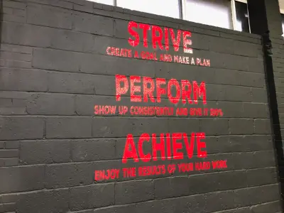 Gym wall vinyl reading Strive, Perform, Achieve in red on grey brick
