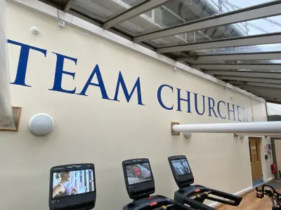 Gym wall with red ship logo and blue Team Churcher's lettering above treadmills