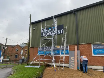 Installer fitting an Athlete Movement wall sign on a gym building using scaffold and ladders